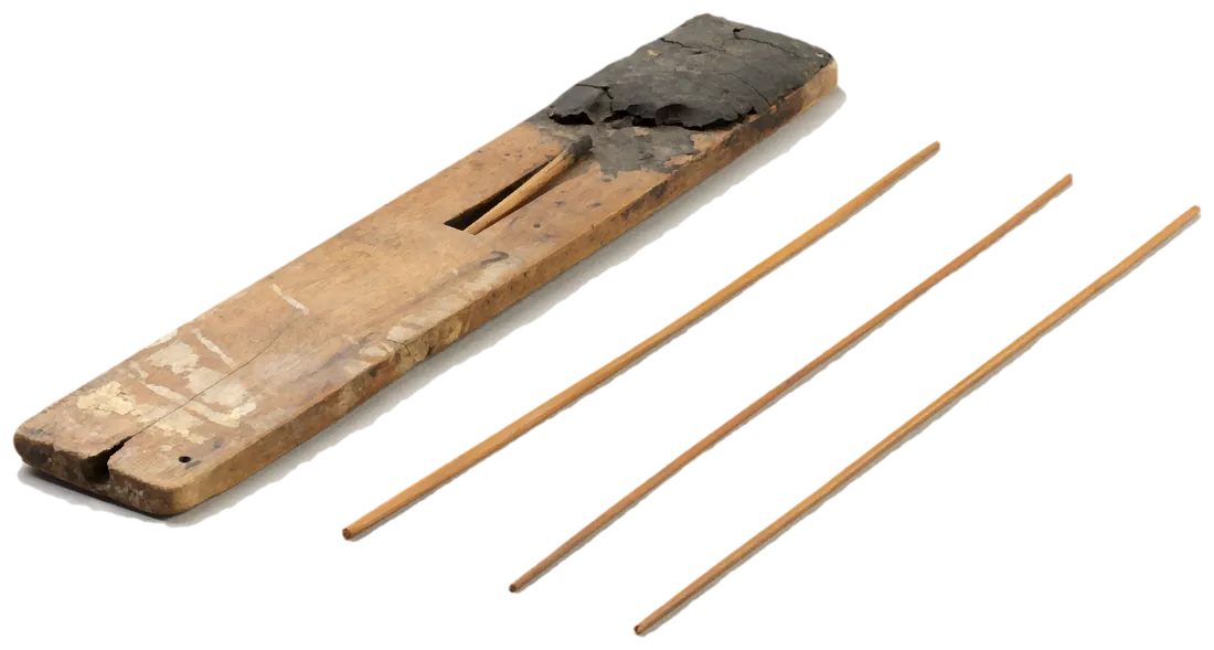 A scribe’s wooden palette with its black ink cake, and three reed pens lying beside it.